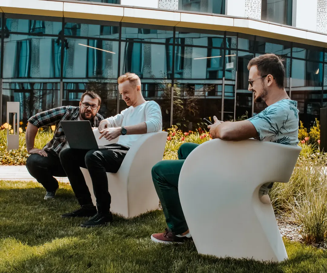Três homens sentam-se em cadeiras brancas modernas do lado de fora de um prédio, trabalham juntos num computador portátil. Eles parecem relaxados e concentrados, com um deles a sorrir enquanto olha para o seu tradutor. Relva verdejante e flores rodeiam-nos.