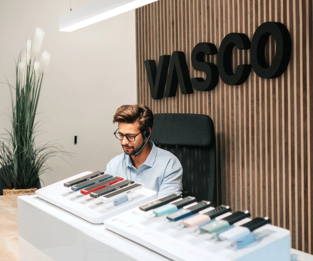 Um homem do serviço ao cliente senta numa secretária exibindo vários dispositivos Vasco Translator em várias cores, com uma grande placa 'VASCO' na parede de madeira atrás dele.