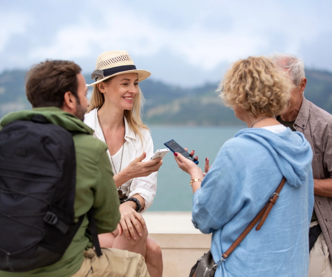 Quatro adultos, três visíveis e um parcialmente obscurecido, conversam ao ar livre junto à água. Duas mulheres, uma delas sorridente e de chapéu, mostram algo nos seus tradutores que seguram nas mãos para o grupo.