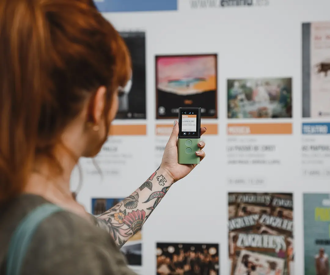 Uma pessoa de cabelo ruivo e com uma tatuagem floral no braço segura um dispositivo digital verde, apontando-o para um quadro com vários cartazes e informações sobre o evento.