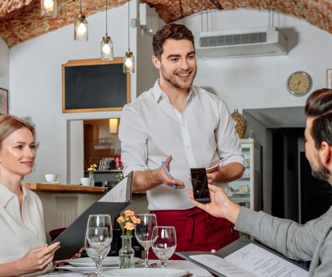 Um empregado de mesa sorridente está à mesa de um restaurante, enquanto um cliente sentado segura um tradutor na mão. Uma cliente do sexo feminino senta-se perto, segurando um menu. A mesa está posta com copos, pratos e uma pequena jarra de flores.