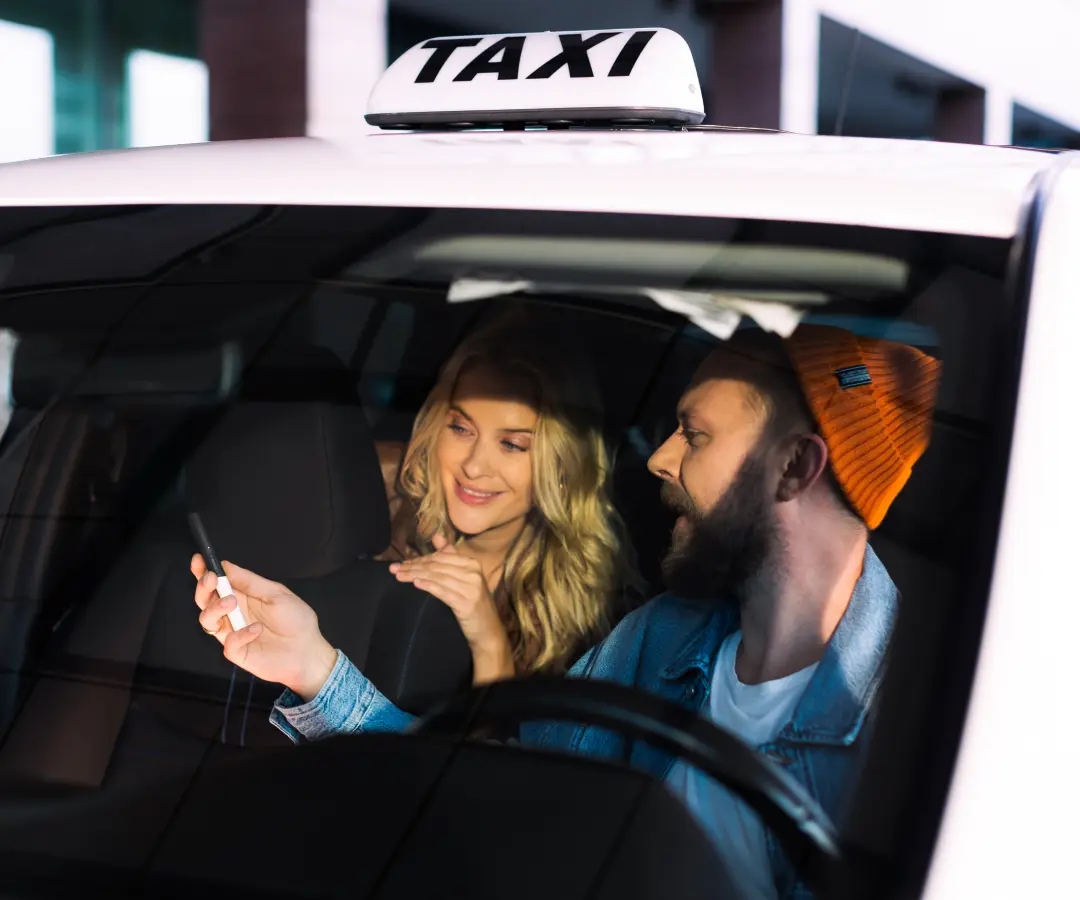 Um homem senta-se no banco do condutor de um táxi e mostra algo a uma mulher no banco de trás através de um tradutor que tem na mão. A mulher sorri com interesse. Vê um sinal de táxi no tejadilho do carro.