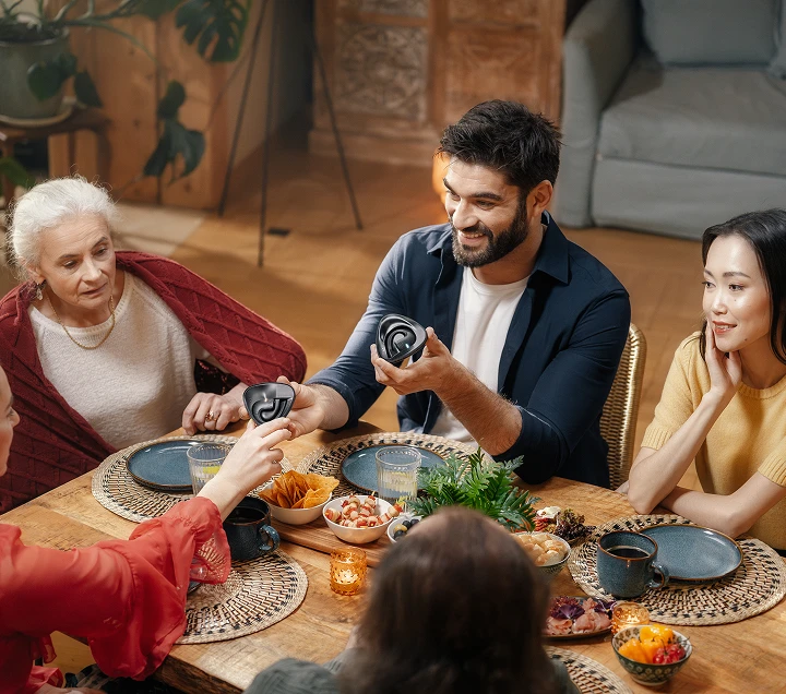 Pessoas de várias nacionalidades reuniram-se à volta de uma mesa, saboreando uma refeição e participando em discussões animadas, criando um ambiente acolhedor e convidativo enquanto trocavam entre si o auricular Vasco Translator E1.