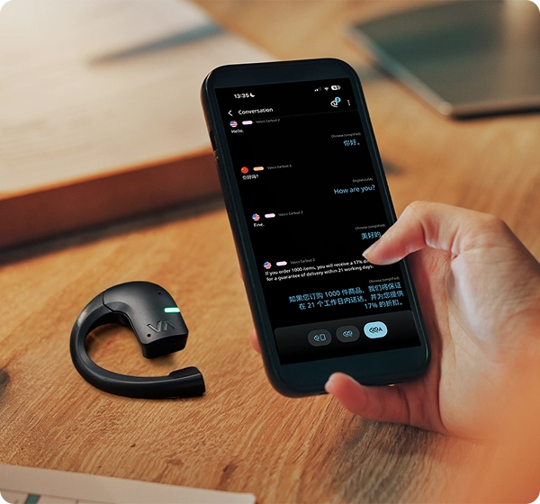 Grande plano de uma mão a segurar um smartphone que exibe uma conversa com tradução em tempo real entre inglês e chinês. Ao lado do telemóvel, um auricular de tradução Vasco preto com um indicador LED verde brilhante repousa sobre uma secretária de madeira. 
