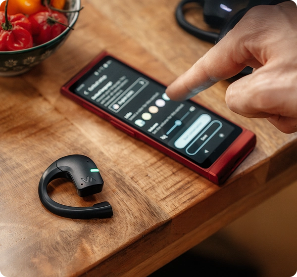 Um grande plano do dedo de uma pessoa a ajustar preferências como a cor e o volume no ecrã de um dispositivo Vasco Translator V4 vermelho. Ao lado, sobre a mesa de madeira, repousa um auricular preto Vasco Translator E1 com um indicador LED verde a brilhar, com uma taça de pimentos vermelhos visível ao fundo. 