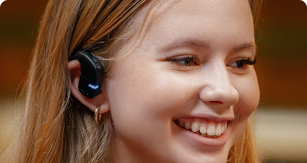 Um grande plano do perfil de uma jovem sorridente a usar um auricular Vasco Translation E1 preto. A imagem destaca o design higiénico e de ouvido aberto do dispositivo, mostrando como este assenta confortavelmente na parte exterior da orelha, sem entrar no canal auditivo. 