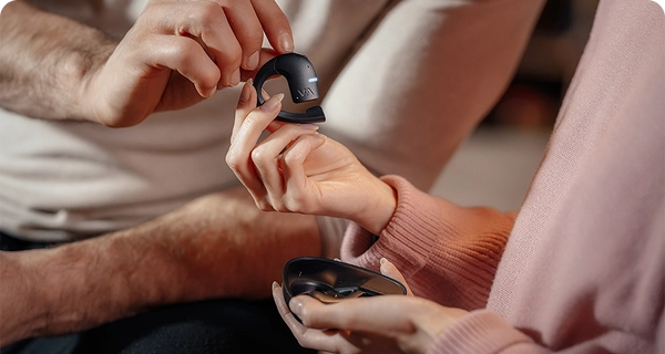 Um grande plano das mãos de uma mulher a segurar delicadamente um auricular Vasco Translation E1 preto entre os dedos, destacando o seu design leve. A mão de um homem toca no dispositivo por cima, enquanto ela segura um estojo de carregamento preto aberto na outra mão. 