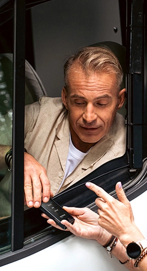 Um homem sentado num carro, a falar com o dispositivo Vasco Q1 que alguém lhe está a entregar.