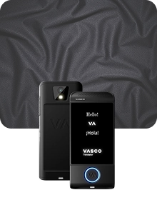 Uma imagem de apresentação do produto que mostra um dispositivo Vasco Translator preto sob dois ângulos, tendo como pano de fundo um tecido escuro e texturado. O dispositivo à esquerda apresenta a vista traseira, destacando a lente da câmara com flash e o logótipo «VA» em relevo. O dispositivo à direita mostra a vista frontal com o ecrã iluminado, exibindo as saudações «Hello!», «VA», «¡Hola!» e o logótipo «VASCO Translator». Um botão circular na parte inferior da frente do dispositivo está contornado por uma luz azul brilhante. O fundo é constituído por tecido cinzento-carvão drapeado com uma textura subtil semelhante a malha.