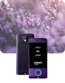Uma imagem de apresentação do produto que mostra um dispositivo Vasco Translator roxo sob dois ângulos, tendo como pano de fundo um tecido escuro e texturado. O dispositivo à esquerda apresenta a vista traseira, destacando a lente da câmara com flash e o logótipo «VA» em relevo. O dispositivo à direita mostra a vista frontal com o ecrã iluminado, exibindo as saudações «Hello!», «VA», «¡Hola!» e o logótipo «VASCO Translator». Um botão circular na parte inferior da frente do dispositivo está contornado por uma luz azul brilhante. A composição tem como pano de fundo um cenário onírico e desfocado de flores roxas de urze banhadas por uma luz solar suave.