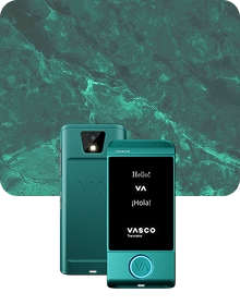 Uma imagem de apresentação do produto que mostra um dispositivo Vasco Translator turquesa sob dois ângulos, tendo como pano de fundo um tecido escuro e texturado. O dispositivo à esquerda apresenta a vista traseira, destacando a lente da câmara com flash e o logótipo «VA» em relevo. O dispositivo à direita mostra a vista frontal com o ecrã iluminado, exibindo as saudações «Hello!», «VA», «¡Hola!» e o logótipo «VASCO Translator». Um botão circular na parte inferior da frente do dispositivo está contornado por uma luz azul brilhante. O fundo apresenta um luxuoso padrão de mármore verde com veios de cor clara.