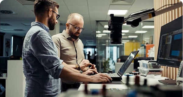 Dois homens com barba e óculos trabalham juntos numa secretária alta, num escritório ou laboratório moderno. Um deles, com uma camisa azul-clara, está a usar um computador portátil, enquanto o outro, com uma camisa bege, está ao seu lado, apontando para um pequeno componente eletrónico que tem na mão. A secretária está equipada com um monitor de grandes dimensões, várias ferramentas eletrónicas e uma câmara montada num braço ajustável. O fundo mostra um espaço de trabalho luminoso e em plano aberto, com divisórias de vidro. 