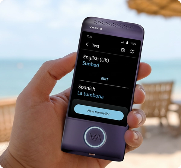 Um plano de perto de uma mão a segurar um tradutor eletrónico Vasco de cor roxa. O ecrã exibe a tradução de um texto do inglês (Reino Unido) para o espanhol, mostrando a palavra «Sunbed» traduzida como «La tumbona». O fundo é uma cena de praia ensolarada e luminosa, com uma imagem difusa do oceano, areia dourada, várias espreguiçadeiras e um guarda-sol de palha. 