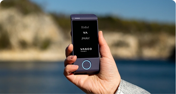 Um grande plano de uma mão a segurar um tradutor eletrónico Vasco roxo, com um fundo exterior luminoso. O ecrã exibe as saudações «Hello!», «¡Hola!» e o logótipo «VASCO Translator». O fundo apresenta uma imagem com efeito de desfocagem suave de uma paisagem costeira ensolarada, com águas azuis e falésias rochosas de cor clara sob um céu límpido. A pessoa que segura o dispositivo usa uma manga de casaco cinzenta clara ou prateada com textura. 