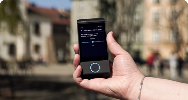 Um grande plano da mão de uma pessoa a segurar um tradutor eletrónico Vasco preto, tendo como pano de fundo desfocado uma praça urbana com edifícios históricos. O ecrã do dispositivo exibe as definições de «Velocidade de leitura da tradução», com um cursor ajustado para «Normal» e uma nota que diz: «Ajuste a velocidade de leitura da tradução movendo o cursor.» A pessoa que segura o dispositivo usa uma pulseira prateada. A luz do dia intensa destaca a interface do ecrã, tornando as definições fáceis de ver. 