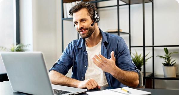Um homem de aparência simpática, com barba e cabelo escuro, está sentado à secretária, usando auscultadores profissionais com microfone. Ele sorri e gesticula com uma mão enquanto olha para o ecrã de um computador portátil, parecendo estar no meio de uma videochamada ou a prestar assistência técnica. Veste uma camisa azul de botões por cima de uma t-shirt branca. Sobre a secretária à sua frente encontram-se um computador portátil, um bloco de notas e uma caneta amarela. O fundo é um espaço de escritório luminoso e moderno, com uma prateleira minimalista de metal preto e vasos de plantas verdes. 