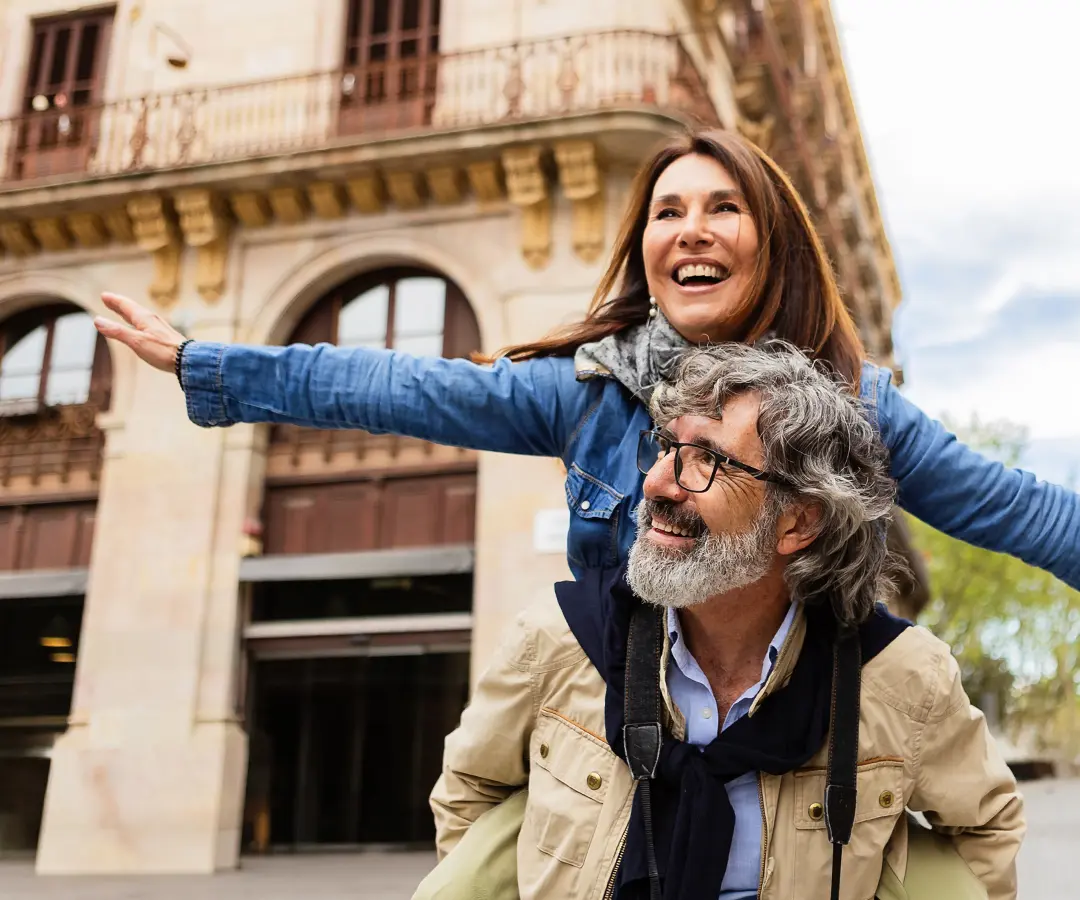 Um senhor de idade, com um ar alegre, leva uma mulher às costas numa rua de uma cidade europeia; ambos sorriem de orelha a orelha enquanto ela aponta com o braço estendido para o horizonte. Ele usa óculos, um casaco bege e tem uma máquina fotográfica; ela usa um casaco de ganga e um lenço. 
