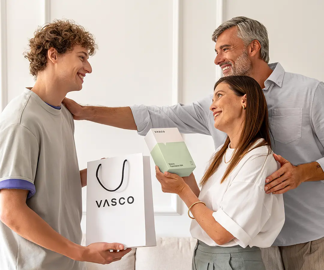 Uma cena comovente em que três pessoas sorriem juntas. À esquerda, um jovem de cabelo encaracolado segura um saco de compras branco com a inscrição «VASCO». No centro, uma mulher segura uma caixa branca e verde-clara com a inscrição «Vasco Translator M4», olhando alegremente para o jovem. Atrás dela, um homem de cabelos grisalhos sorri calorosamente, com uma mão no ombro da mulher e a outra no ombro do jovem. Estão numa sala luminosa com uma parede de cor neutra e um sofá ao fundo. 