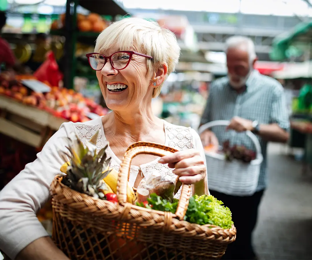 Uma senhora de idade, sorridente, com cabelo loiro curto e óculos vermelhos, carrega um cesto de vime cheio de legumes e fruta fresca num mercado ao ar livre. No fundo desfocado, outra pessoa faz compras entre as bancas de produtos hortícolas.